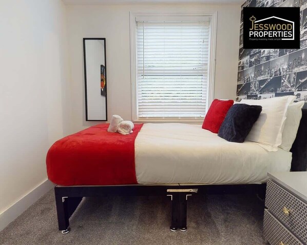 A neatly arranged bedroom with a bed, a window with blinds, a framed picture on the wall, and a logo on the wall. - Contractors 3 Bed |WiFi | Sleeps 5 Long Term Stays (Luton)