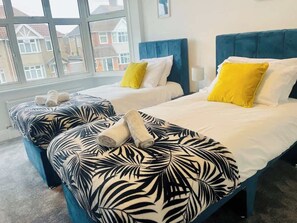 A hotel room with two beds, each with a blue headboard, white bedding, and a yellow pillow. There is a window with a view of houses outside. - Contractor Friendly | 2 Bath |Free Parking | Wifi (Central Bedfordshire)