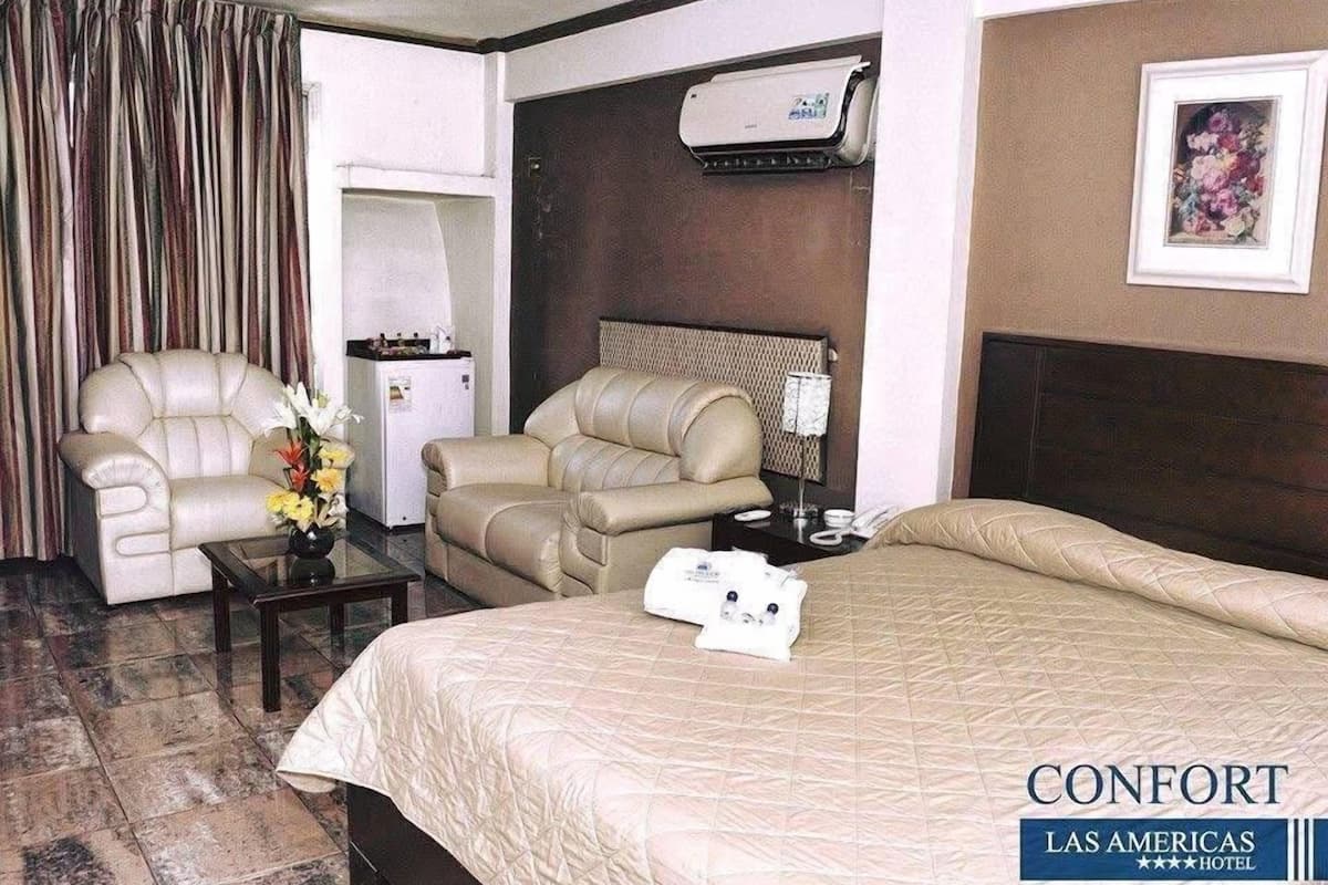 A hotel room with a bed, two sofas, a desk, a refrigerator, an air conditioning unit, and a painting on the wall.