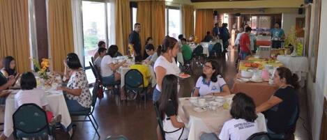 Uma sala de jantar com mesas e cadeiras, pessoas refeiscando e um buffet ao fundo.