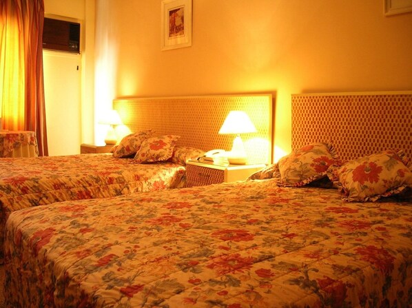 A hotel room with two beds, floral bedspreads, a headboard, a nightstand with a lamp, and a framed picture on the wall. - Hotel Las Américas (Santa Cruz)