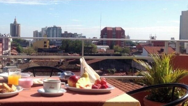 A balcony with a table set for breakfast, including a plate of fruit, a glass of orange juice, and a cup. The view includes a cityscape with buildings and a clear sky.