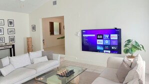 Ein Wohnzimmer mit einem weißen Sofa, einem Glastisch und einem an der Wand befestigten Flachbildfernseher, der eine Roku-Oberfläche anzeigt.