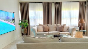 Ein Wohnzimmer mit einem beigen Sofa, einem Couchtisch mit Schachfiguren, einem Fernseher, der eine Strandansicht zeigt, und einer Zimmerpflanze.