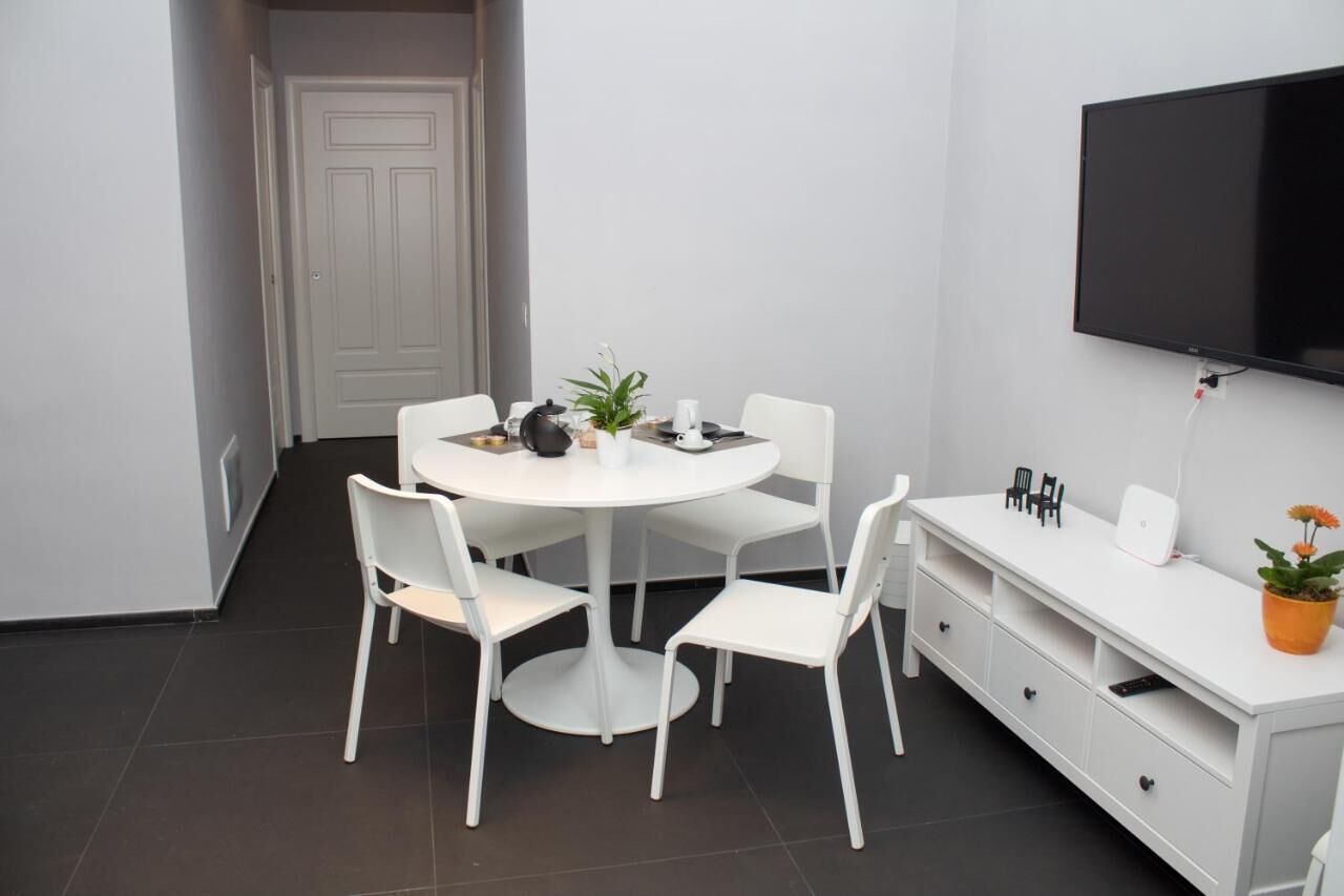 A modern, minimalist room with a round dining table, white chairs, a flat-screen TV mounted on the wall, and a white console table with drawers and a potted plant.