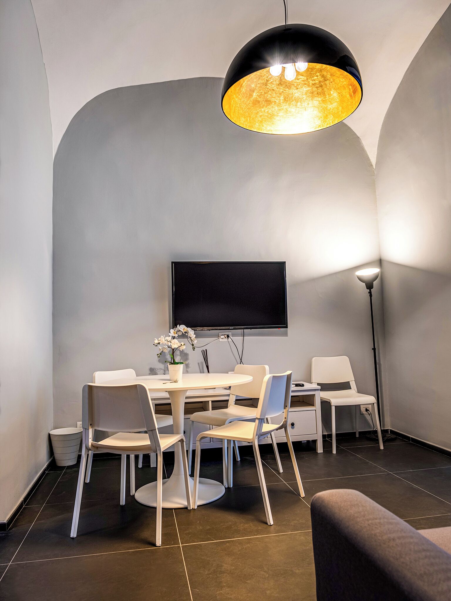 A modern dining area with a round table, white chairs, a black ceiling light, and a flat-screen TV mounted on the wall.