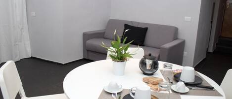 A dining area with a round table set for two, a grey sofa in the background, and a potted plant on the table.