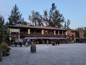 Um pátio com chão de pedra, rodeado por edifícios com arquitetura tradicional, incluindo um restaurante com mesas ao ar livre.