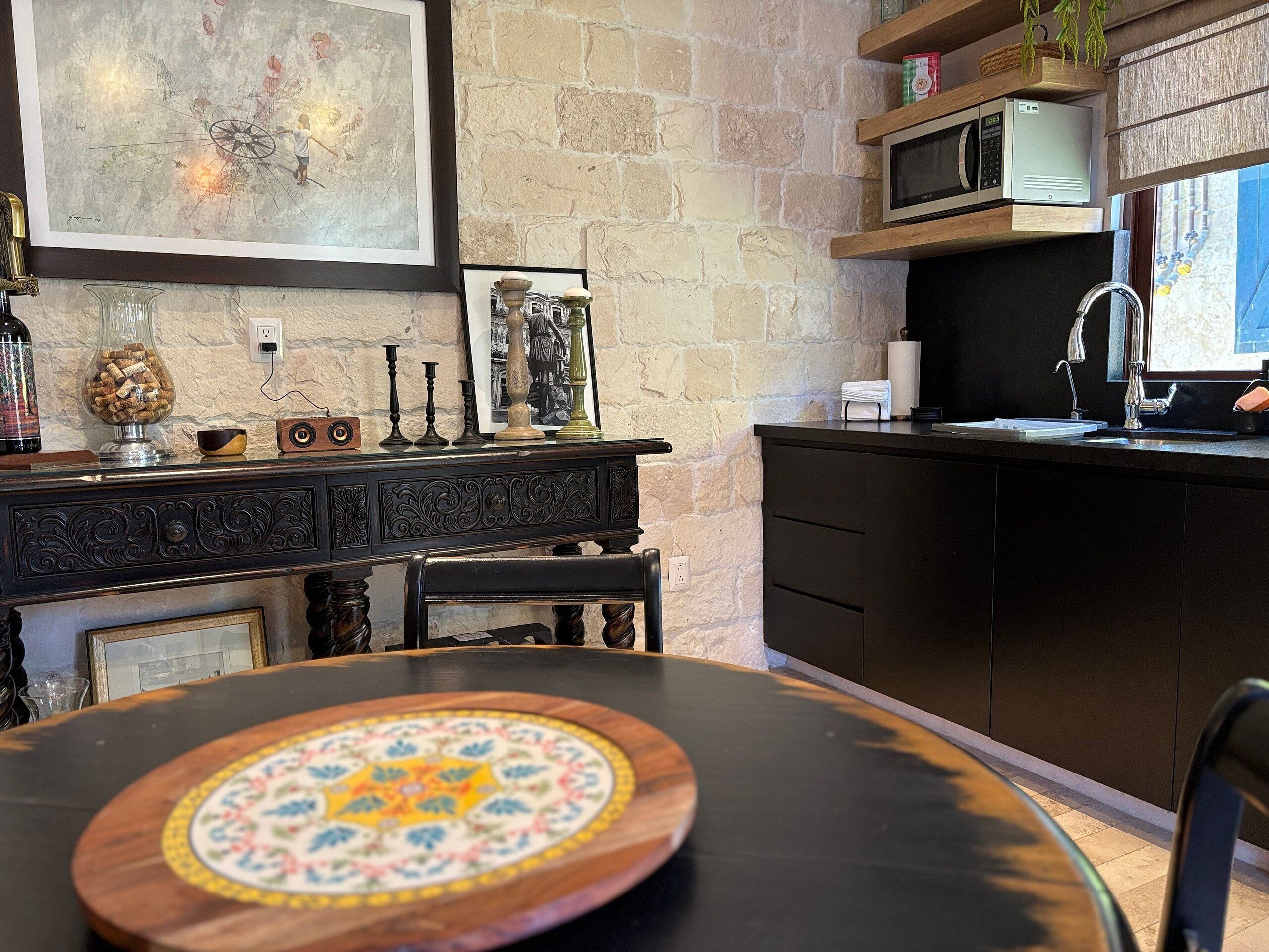 A kitchen with a stone wall, a wooden dining table with a decorative plate, a black countertop, and a microwave above the sink.