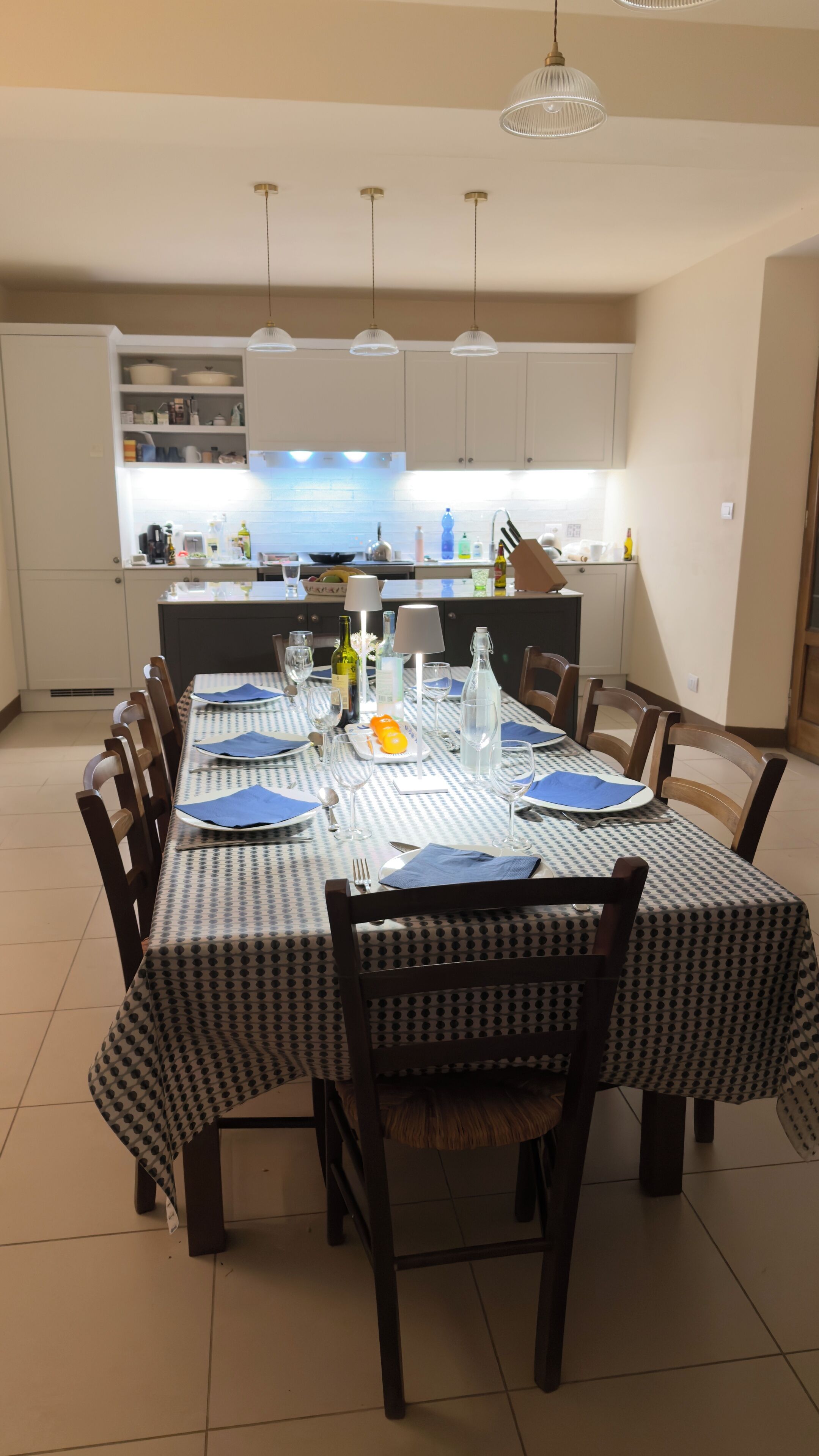 A dining area with a table set for four, a kitchen in the background, and pendant lights above.