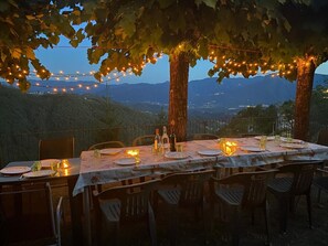 An outdoor dining setup with a table set for four, surrounded by chairs, under a canopy of trees with string lights. - Beautiful family villa with private pool and wonderful mountain views near Barga (Sommocolonia)