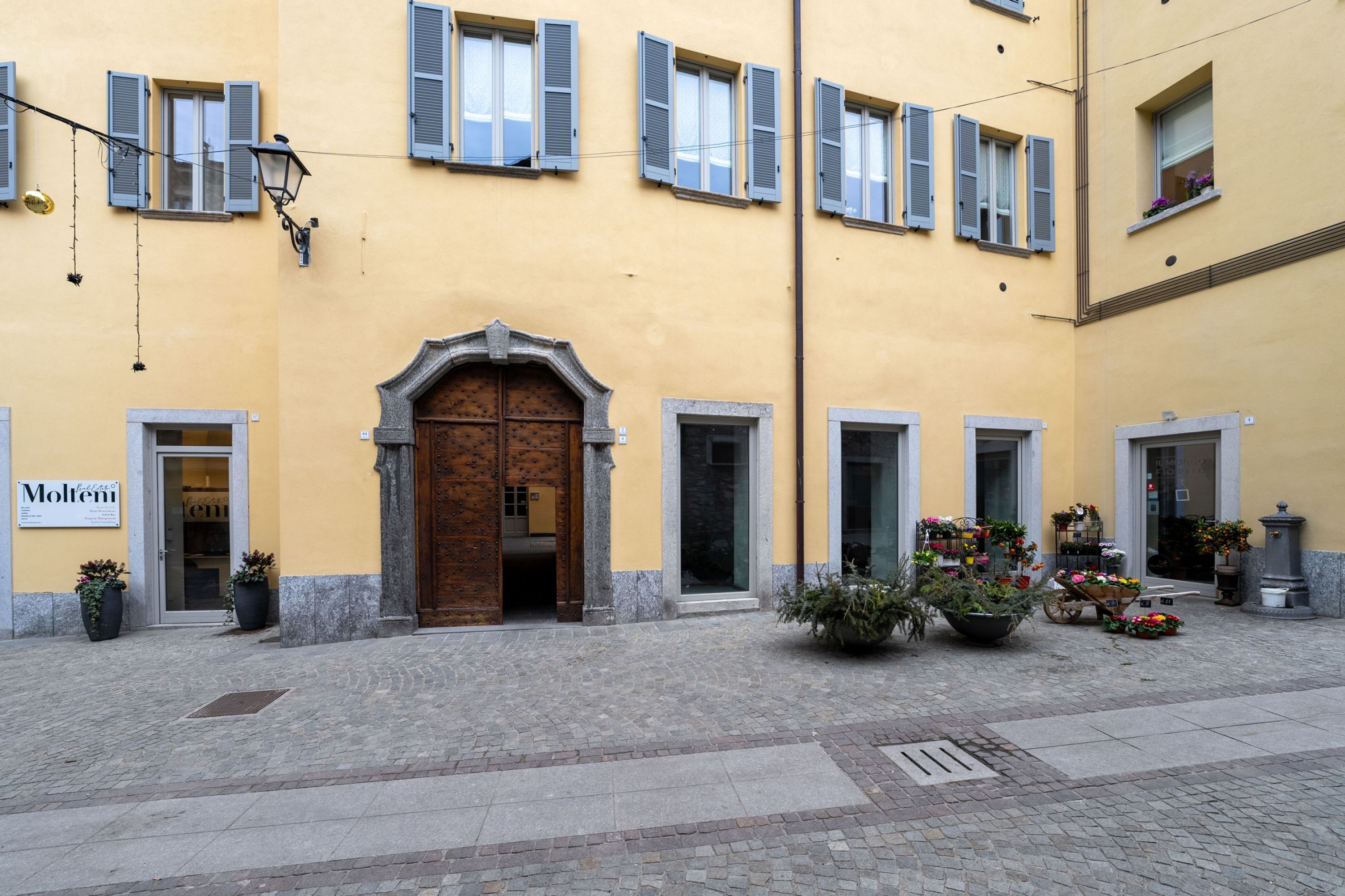 A building with a yellow facade, multiple windows with shutters, a central entrance with a wooden door, and potted plants outside.