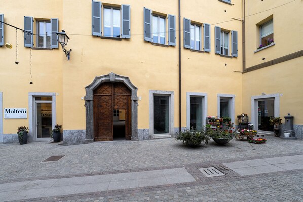 A building with a yellow facade, multiple windows with shutters, a central entrance with a wooden door, and potted plants outside. - Comfy Apartment in Bellano by Wonderful Italy (Bellano)