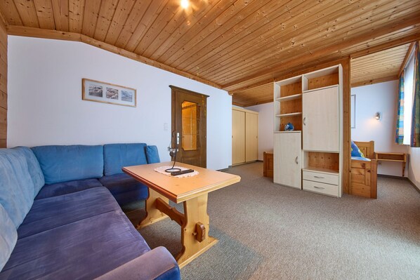 A cozy room with a wooden ceiling, a blue sofa, a wooden table with a book, and a wardrobe with open doors. - Haus Miller (Sankt Veit im Pongau)