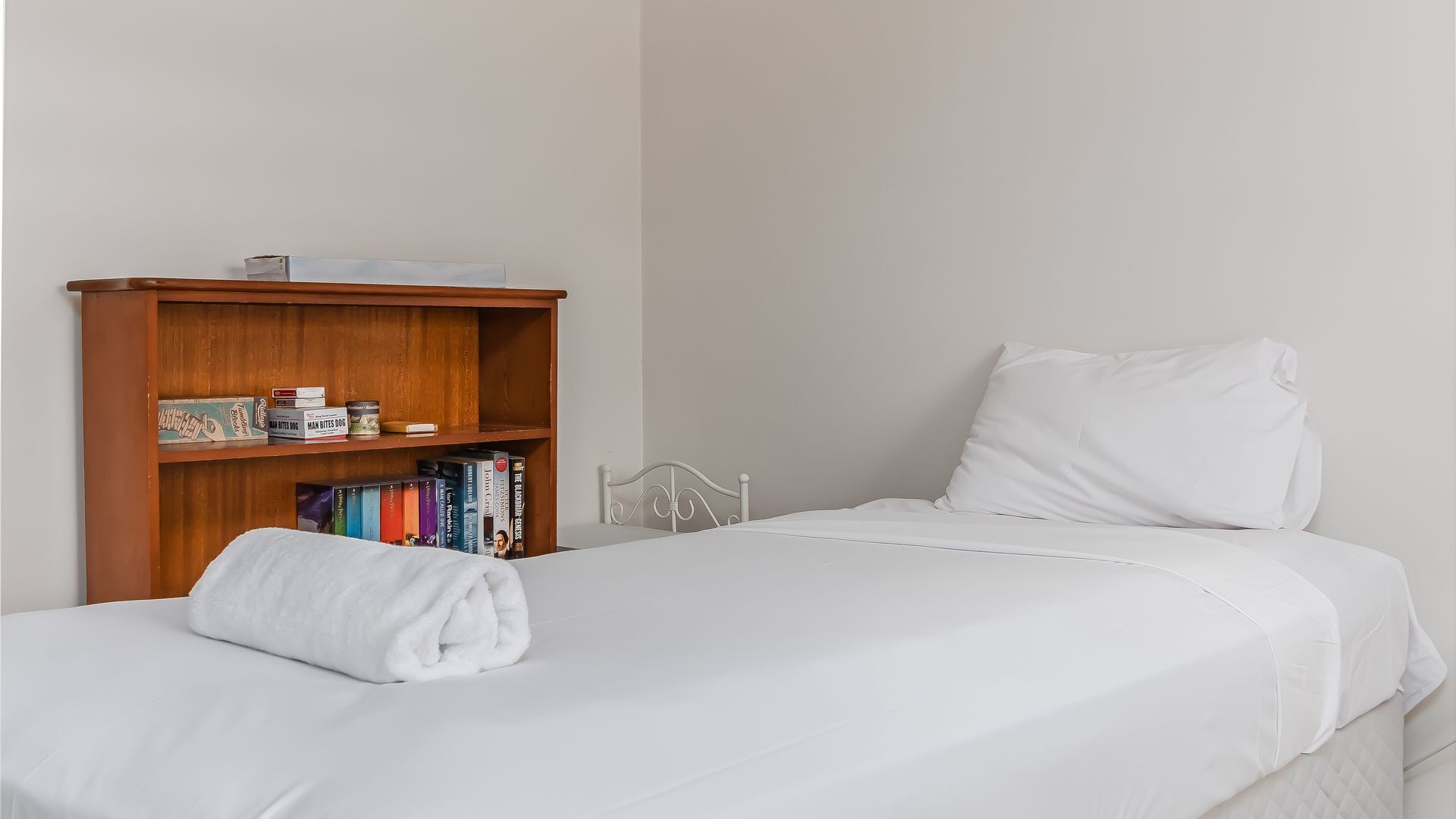 A neatly made bed with white linens, a wooden bookshelf with books and a small box, and a plain wall in the background.