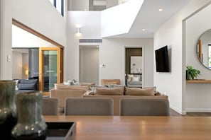A spacious living area with a high ceiling, a dining table, and a TV mounted on the wall.