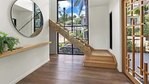 A staircase with wooden steps and metal railings, a round mirror on the wall, and a view of the outdoors through large windows. - Sea Mist—Poolside Oasis near Palm Cove Beachfront (Palm Cove)