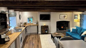 A cozy living room with a fireplace, a blue sofa, and a kitchen area with wooden flooring and white cabinets. - Cosy, comfy & tranquil farm cottage with log fire & country walks from the door (Talgarth)