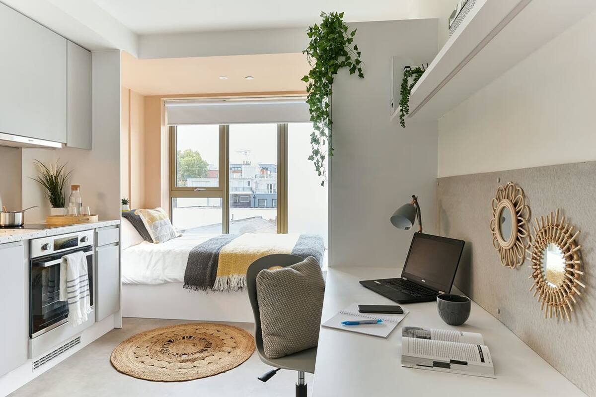 A modern bedroom with a bed, a desk with a laptop, a window, and a kitchen area.
