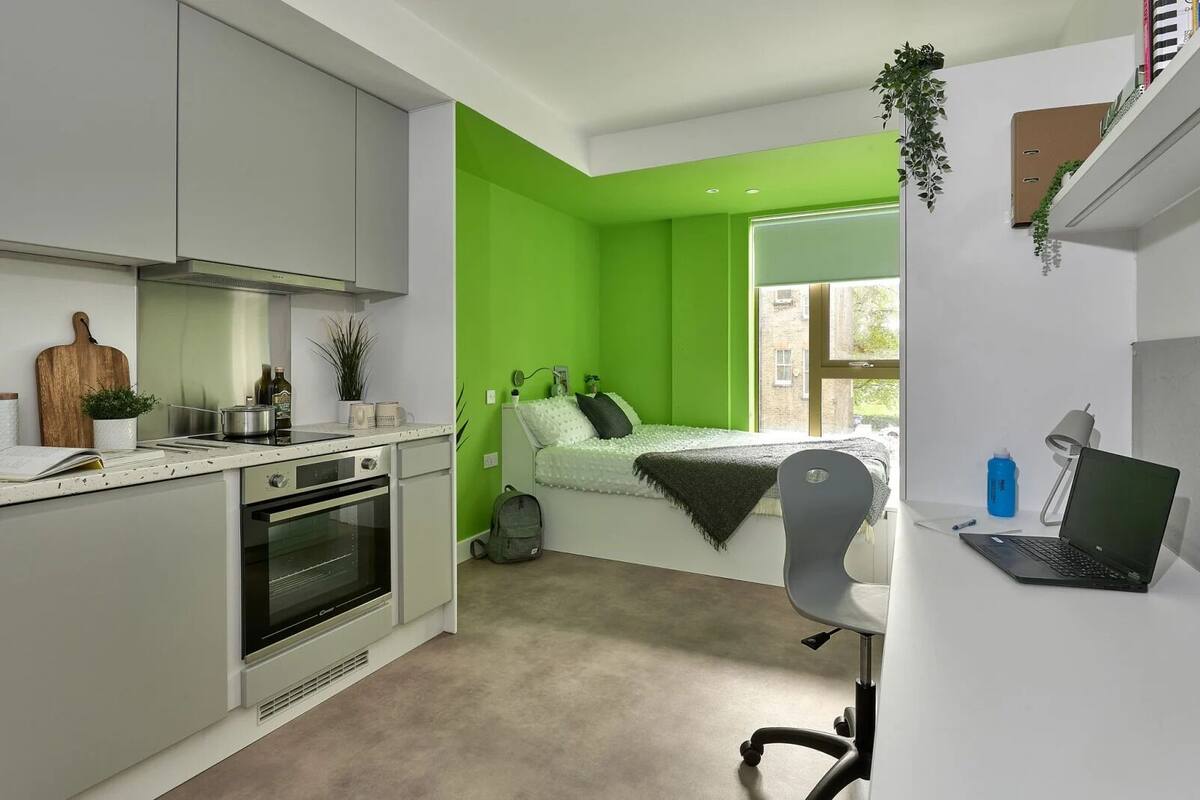 A compact studio apartment with a kitchenette, a bed with green walls, a desk with a laptop, and a window providing a view of the outside.
