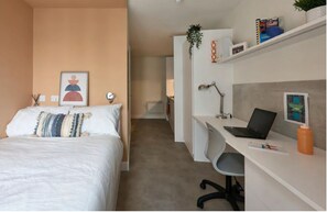 Moderni hotellihuone, jossa on sänky, työpöytä ja tuoli. Työpöydällä on kasvi ja seinällä valokuvakehys.