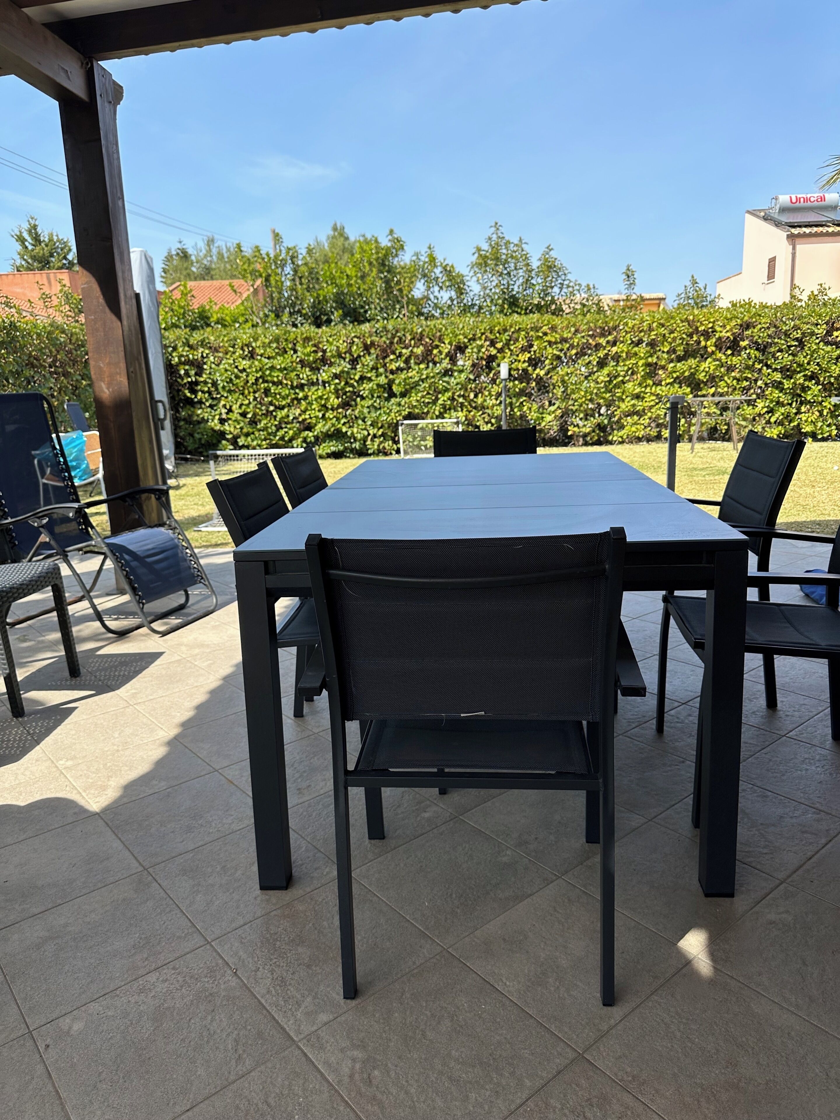 An outdoor patio with a rectangular table and six chairs, surrounded by a tiled floor and a covered area.