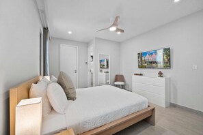 Une chambre à coucher moderne, équipée d’un lit, de tables de chevet, d’un ventilateur de plafond, d’une télévision et d’une œuvre d’art accrochée au mur.