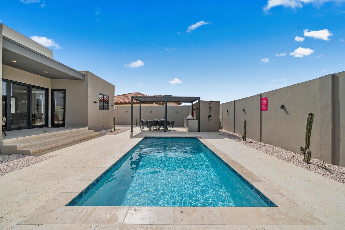 A rectangular swimming pool with a clear blue surface, surrounded by a patio area with steps leading to a covered seating area.