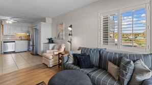 A modern kitchen with white cabinets, a built-in oven, and a dishwasher. There is a dining area with a sofa and a table. A large window with blinds provides a view of the outdoors. - A Place At The Beach IV #228 (Myrtle Beach)
