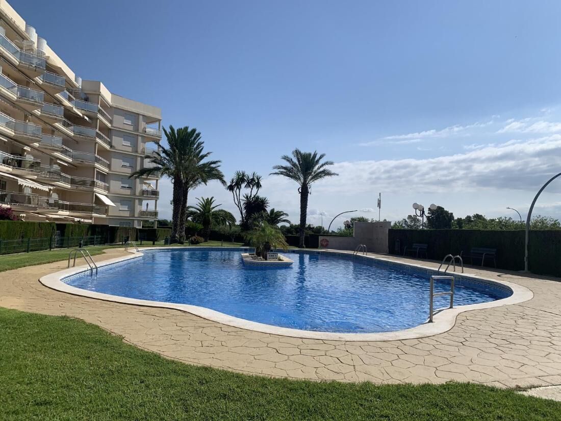Une piscine avec une terrasse carrelée, entourée de palmiers et bordée d’un immeuble à plusieurs étages doté de balcons.