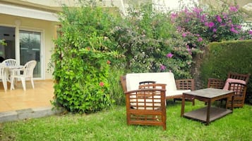 Un patio con mesa y sillas de madera, rodeado de abundante vegetación y plantas con flores.