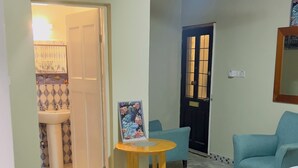 Una stanza con un letto, una sedia, un piccolo tavolo con un quadro, una ventola a soffitto e una porta che dà su un'altra stanza.