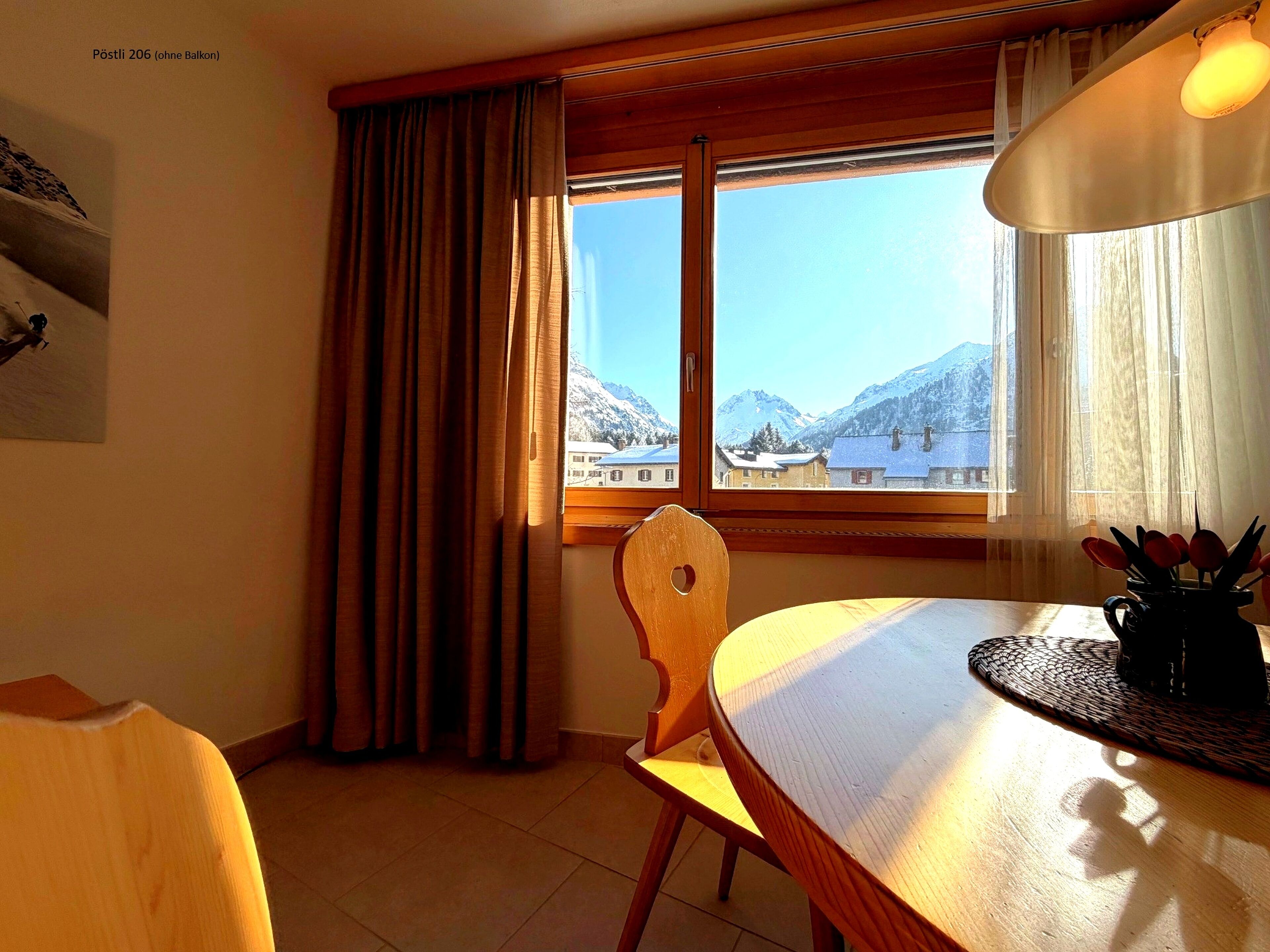 Ein Zimmer mit Holztisch, Stuhl, einem Fenster mit Vorhängen und Blick auf Berge.