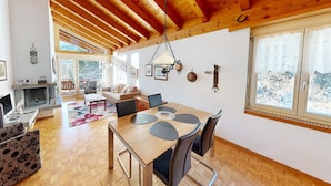A spacious living room with a wooden ceiling, a dining area with a wooden table and chairs, and a fireplace. - Schweizerhaus Apartments (Maloja)