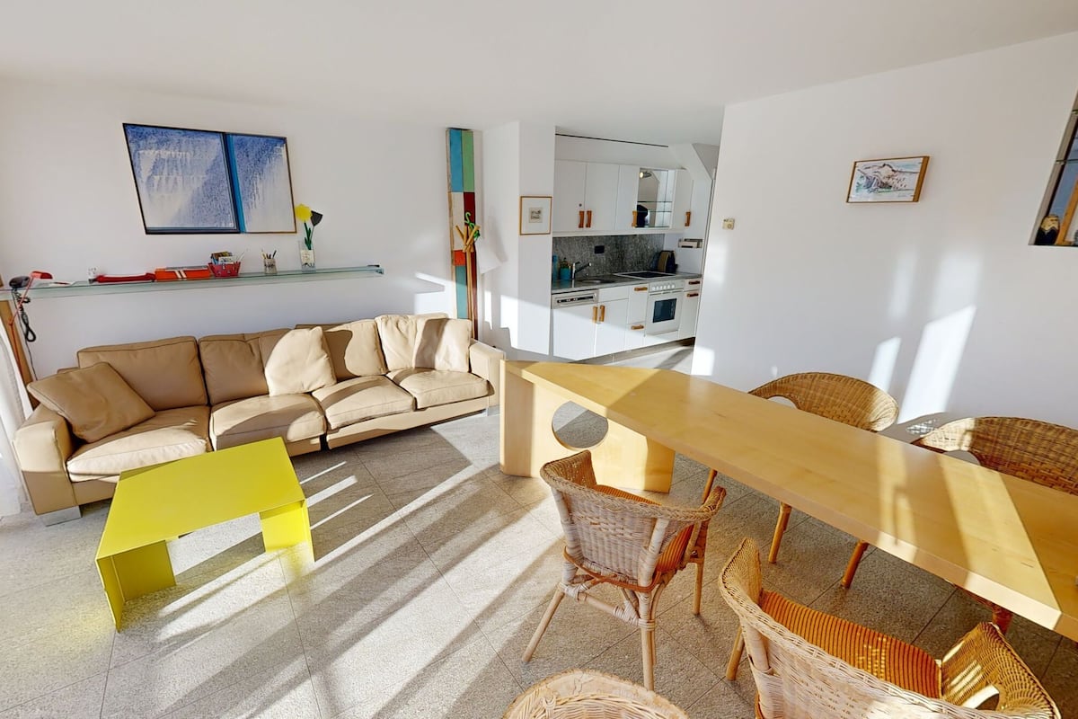 A modern living room with a sofa, a wooden table, wicker chairs, and a kitchen in the background.