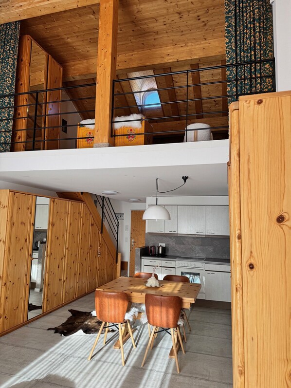 A modern loft-style kitchen with wooden beams, a dining area with leather chairs, and a staircase leading to a lofted sleeping area. - Schweizerhaus Apartments (Maloja)