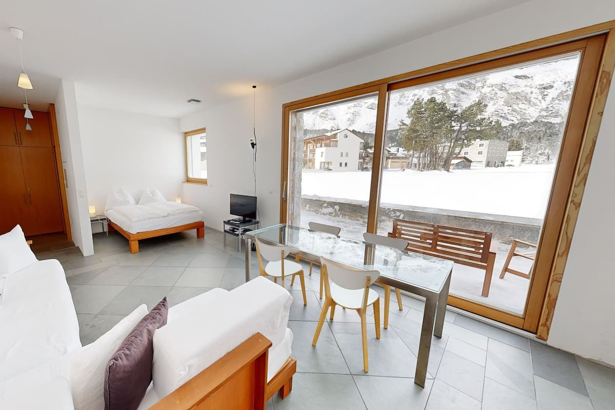 A modern bedroom with a bed, bedside table, and a TV. There is a glass table with chairs in the living area, and a large window with a view of a snowy landscape.