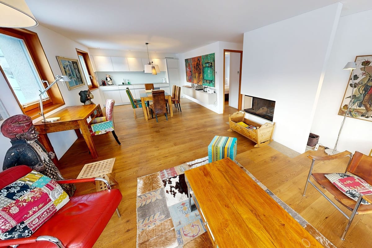 A spacious living room with a wooden floor, a red sofa, a wooden coffee table, and a TV. There is a kitchen area with white cabinets and a dining table with chairs.