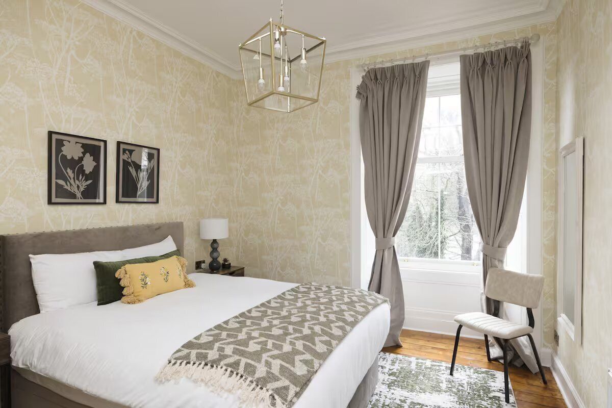 A bedroom with a large bed, a nightstand, a chair, a window with curtains, and a chandelier.