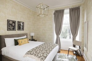 Ein Schlafzimmer mit einem großen Bett, einem Nachttisch, einem Sessel, einem Fenster mit Vorhängen und einer Chandelier.