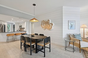 Uma cozinha moderna com armários brancos, uma sala de jantar com mesa e cadeiras redondas, e uma sala de estar com sofá e abajur.