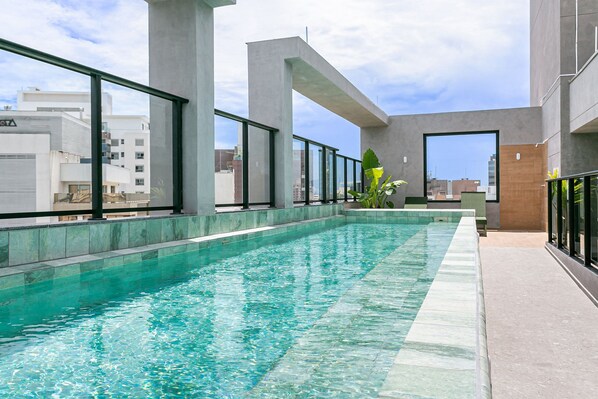 Una piscina en la azotea con agua cristalina, rodeada de amplios ventanales y pilares de hormigón.