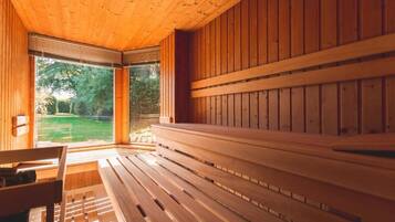 Eine Holzsauna mit Bänken und einer Sitzbank, großen Fenstern und Blick auf eine grüne Außenanlage.