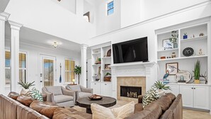 A spacious living room with a large leather sofa, a fireplace, and a television mounted above it. There are white walls and built-in shelves with decorative items. - La Madeleine Coastal Home Resort Pools Quiet Beach (Gulf Shores)