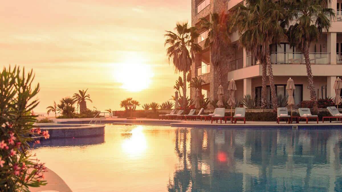 Un espace piscine agrémenté de chaises longues et de palmiers, où se reflète le coucher de soleil.