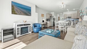 Ein modernes Wohnzimmer mit einer Couch, einem blauen Sessel, einem Essbereich mit weißem Tisch und einer Küche im Hintergrund.