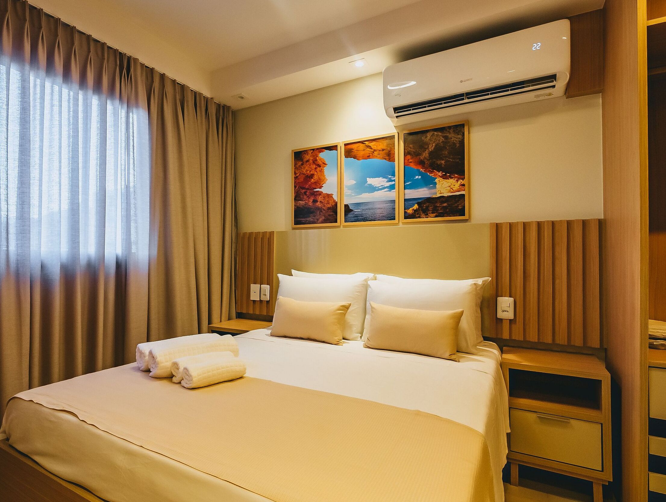 A hotel room with a large bed, beige bedding, a nightstand, a wall-mounted air conditioner, and a window with curtains.