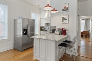 Una cocina moderna con refrigerador de acero inoxidable, muebles blancos, una isla de encimera de mármol y lámparas colgantes rojas.