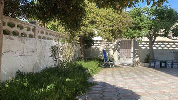 Un cortile lastricato con un sentiero in pietra, un muro bianco con decorazioni intagliate e un albero che offre ombra.