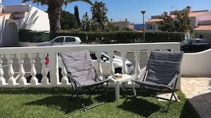 Un espacio al aire libre soleado con dos sillones de descanso y una mesita, rodeado por una cerca blanca y palmeras.